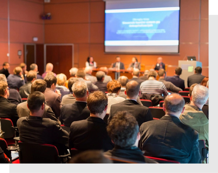 A conference room filled with people seated, facing a panel of speakers at a table and a presentation screen at the front, showcases the latest trends in Sustainable Product Design.